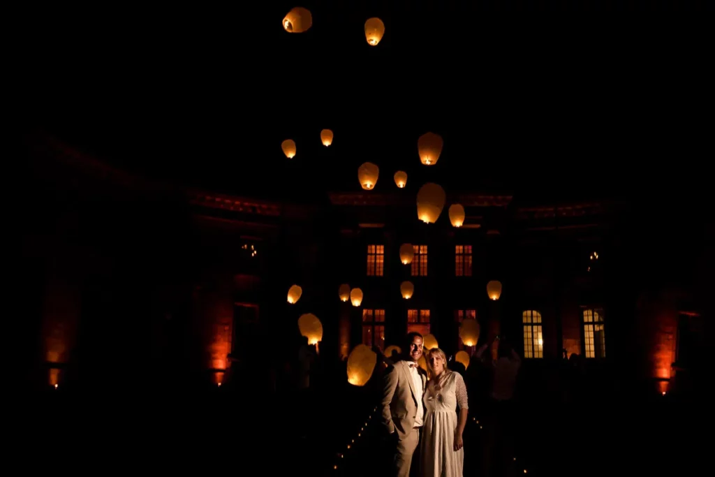 Une mariée et un marié debout devant un bâtiment avec des lanternes dans le ciel.